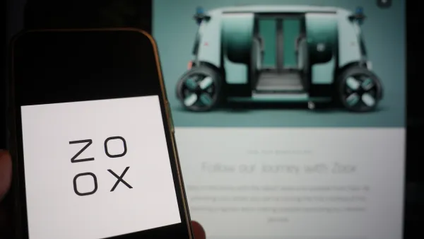 Close-up of smartphone with Zoox logo and screen with blurred image of Zoox autonomous vehicle in background.